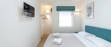 A neatly made bed with a quilted cover and two rolled towels, a window with a blue valance, a framed picture on the wall, and a poster on the wall.