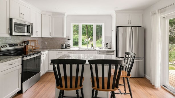 A modern kitchen with white cabinetry, stainless steel appliances, a large island with chairs, and a view of greenery through the window.