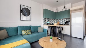 Un soggiorno moderno con un divano color petrolio, un tavolino da caffè rotondo in legno, e una zona cucina con mobili color petrolio e un frigorifero bianco.