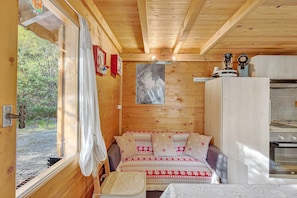 A wooden cabin interior with a bed, a small table, a window with curtains, a kitchen area, and a view of the outdoors. - Chalet 'Les Chalets' with Mountain View, Shared Terrace and Private Garden (Méolans-Revel)