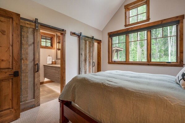 Una camera da letto con un letto grande, porte in legno, una finestra vista alberi e un bagno piccolo con uno specchio e un lavandino.