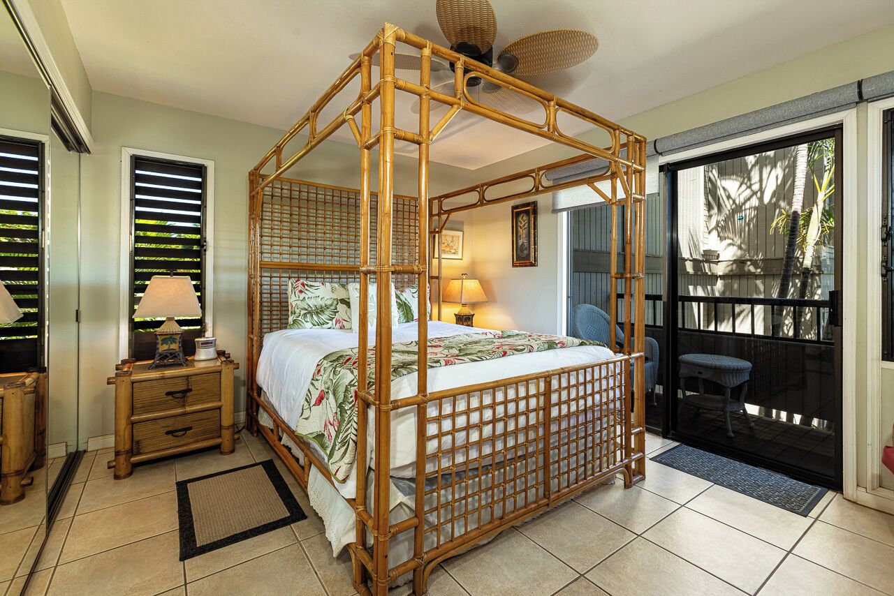 A four-poster bed with a woven canopy, a bedside table with a lamp, a chair, and a view of the outdoors through a sliding door.