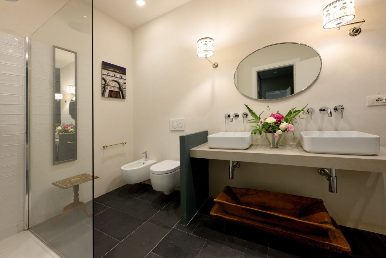 Une salle de bain moderne avec une douche à parois de verre, un bidet, un lavabo blanc avec un miroir et un vase de fleurs.