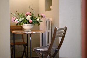 A small round table with a white vase containing pink and white flowers, a wooden chair, and a radiator in the background.