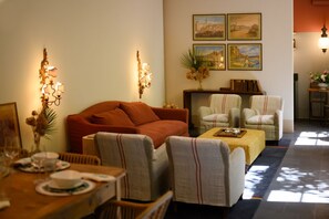 A living room with a red sofa, two armchairs, a coffee table, and a dining table set for four. There are wall-mounted light fixtures and framed pictures on the wall.