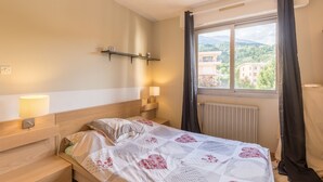 Une chambre à coucher avec un lit, des lampes de chevet, une étagère, une fenêtre avec des rideaux et une vue sur des bâtiments et de la verdure.