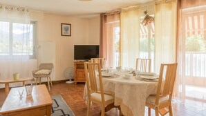 Un coin repas avec une table dressée pour quatre personnes, des chaises en bois, un téléviseur et une fenêtre avec des rideaux.