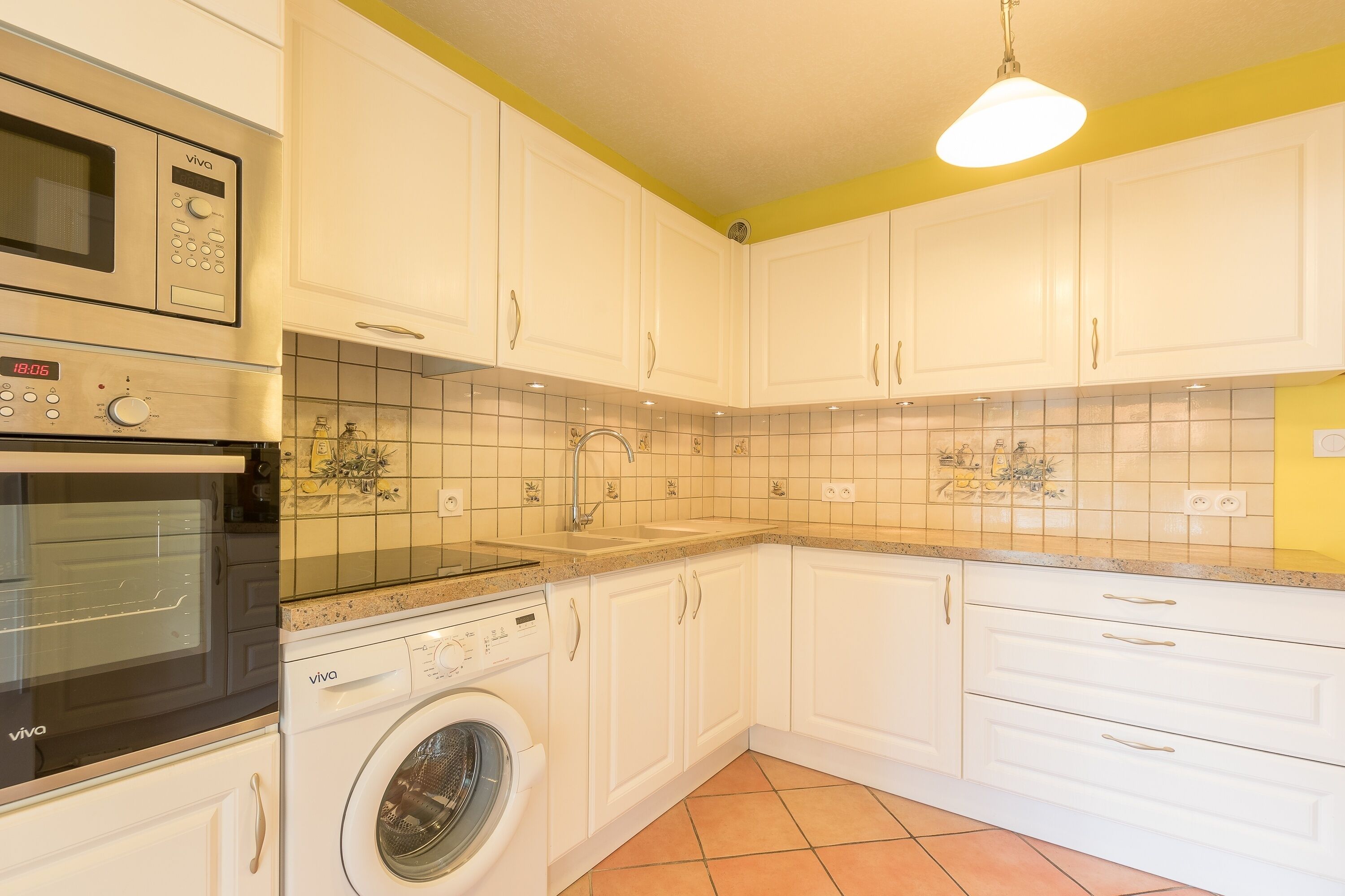 Une cuisine avec des meubles blancs, un four à micro-ondes, un lave-linge et un pare-feu en carrelage.