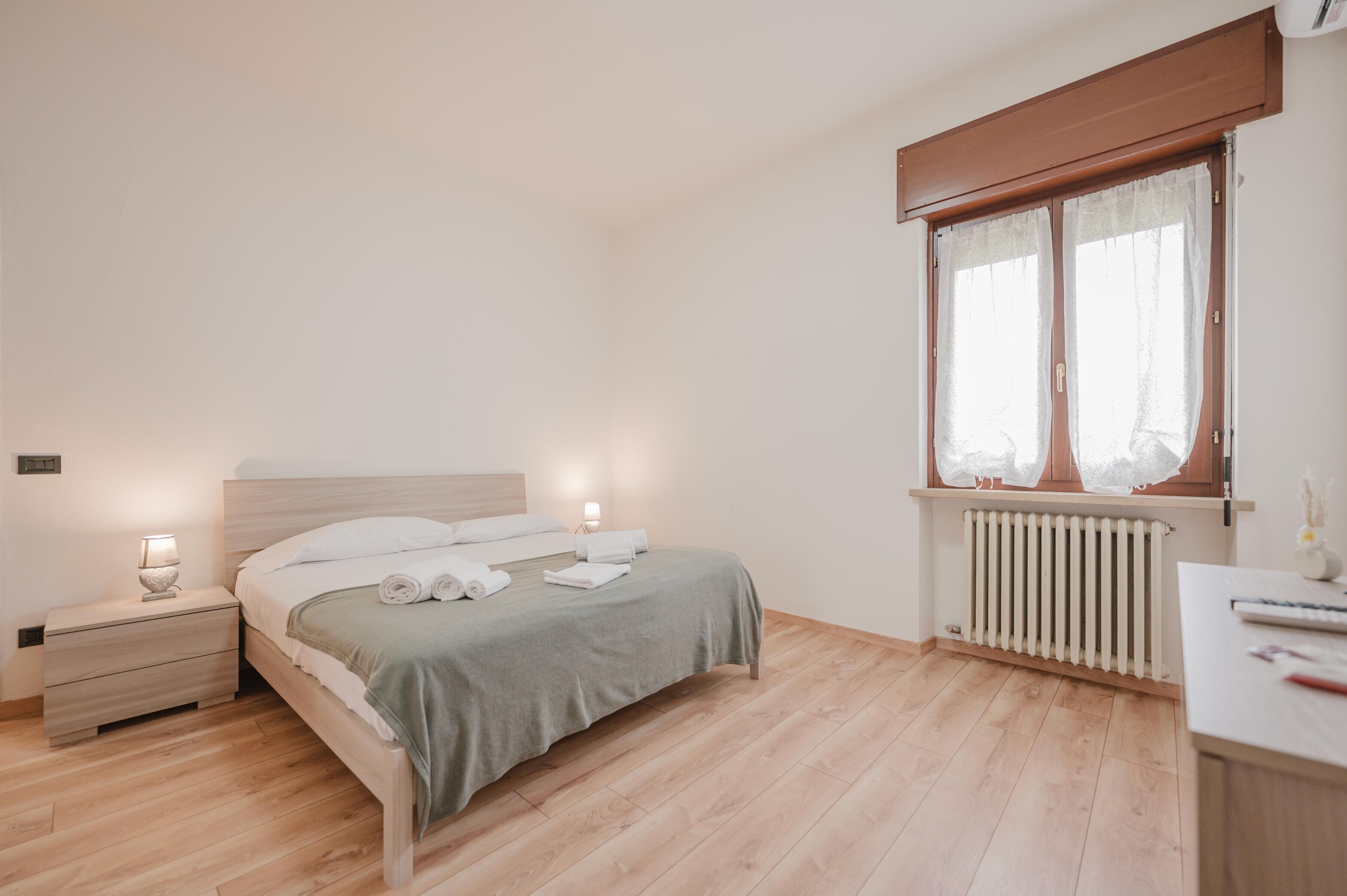 Une chambre à coucher comprenant un lit, des tables de chevet, une fenêtre avec des rideaux, un radiateur et un bureau avec une lampe.