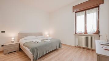 Une chambre à coucher comprenant un lit, des tables de chevet, une fenêtre avec des rideaux, un radiateur et un bureau avec une lampe.