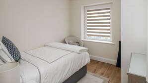Une chambre à coucher avec un lit, une chaise, une fenêtre avec des stores, une lampe et une petite table.