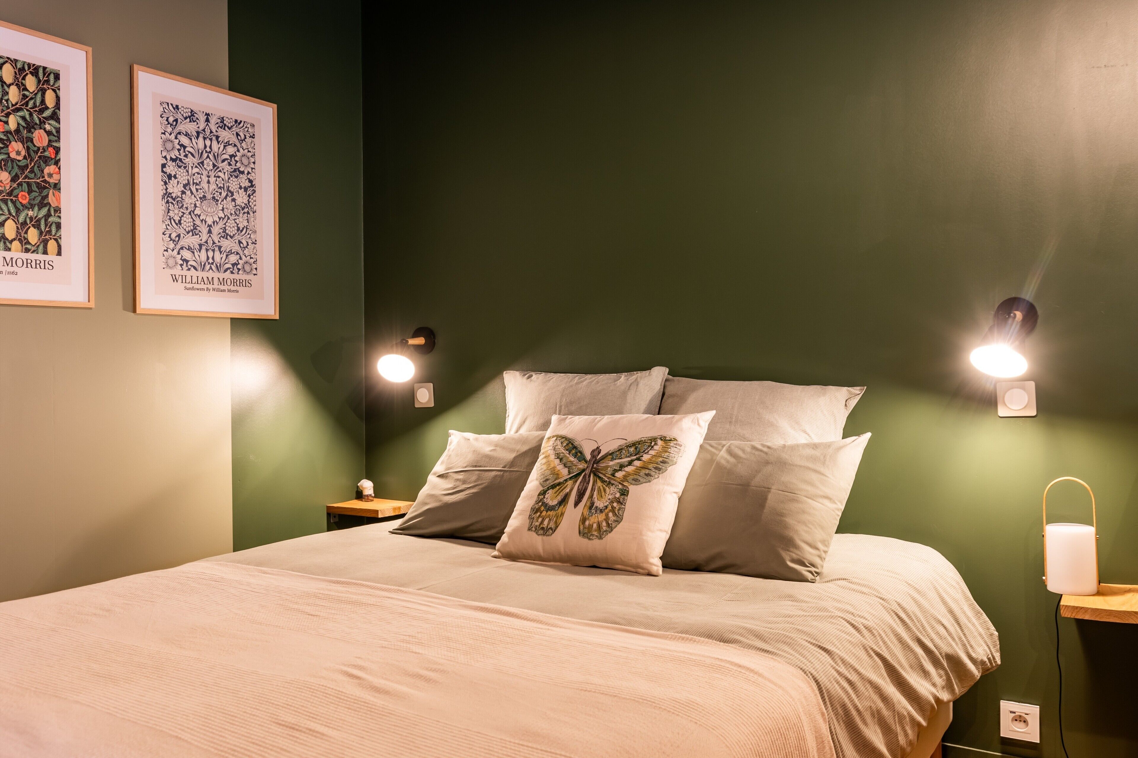 A neatly made bed with a butterfly pillow, two framed artworks on the wall, and wall-mounted reading lights.