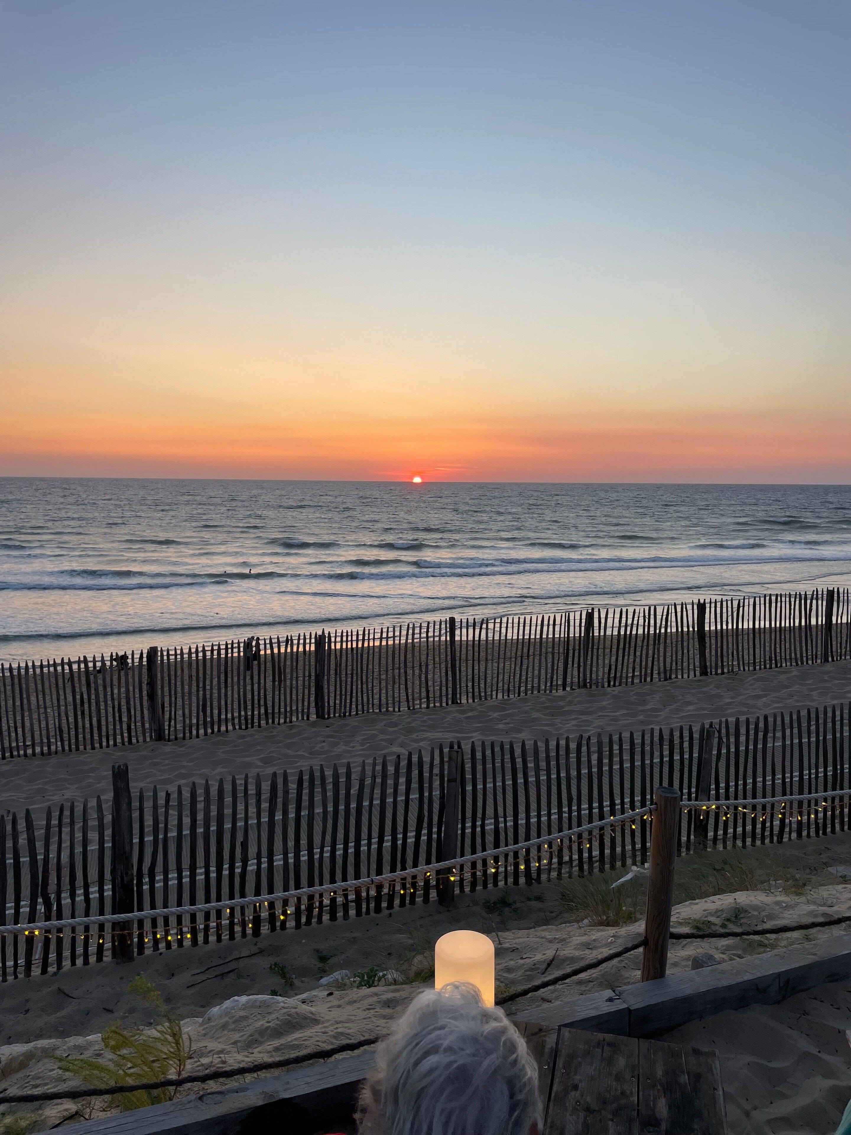 Une vue sur le coucher de soleil au-dessus de l’océan, une clôture en bois et une plage de sable.