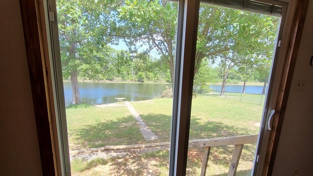 A room with a sliding glass door leading to a backyard with a lawn, a wooden deck, and a view of a lake. - Cozy Lakefront 2-bedroom cabin in tranquil Big Sandy, TX. (Big Sandy)