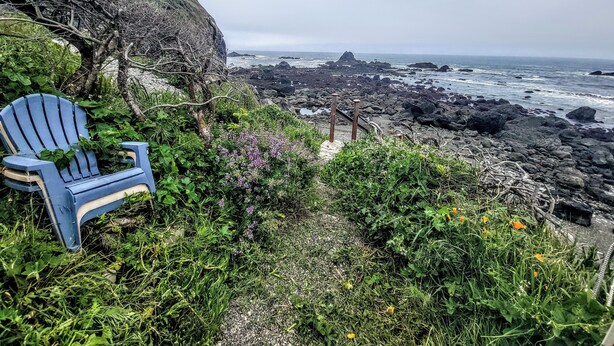 A blue Adirondack chair on a rocky cliff overlooking a rugged coastline with waves crashing against the rocks.