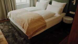 A neatly made bed with white linens and a brown throw blanket, a bedside table with a lamp, and a view of the street through a window with curtains.