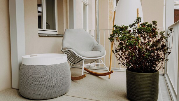 A balcony with a white rocking chair, a woven ottoman, and a potted plant.