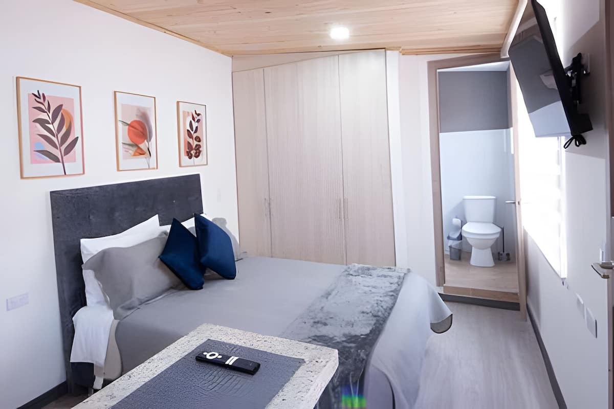 A compact hotel room with a bed, a bedside table with a remote control, a wardrobe, and a bathroom visible through an open door.