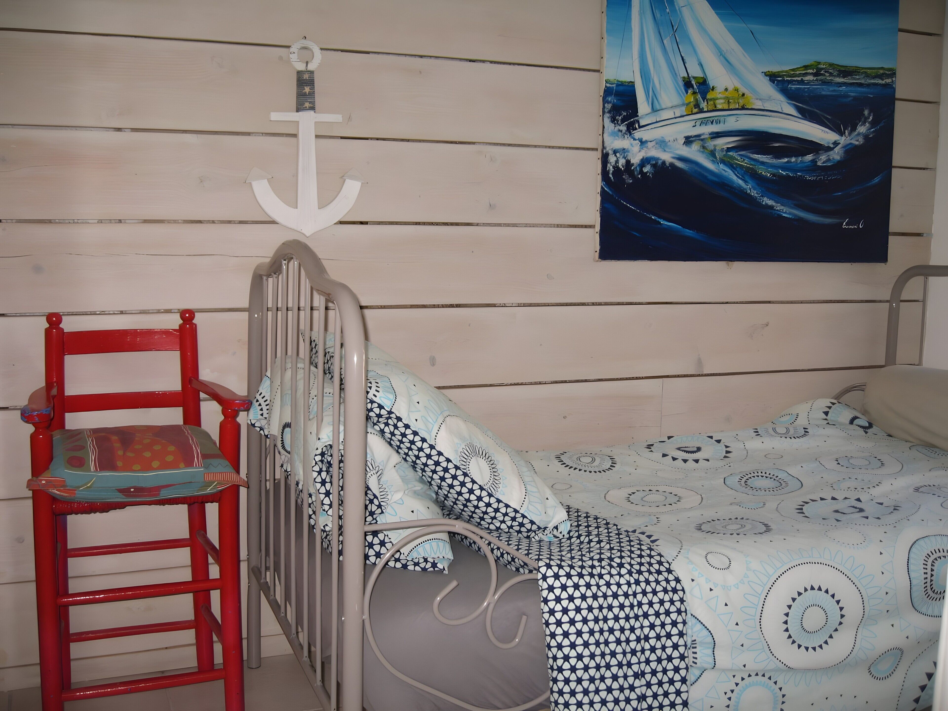 Une pièce avec un berceau en métal, une chaise rouge, une couvre-lit à motifs et un tableau représentant un voilier accroché au mur.