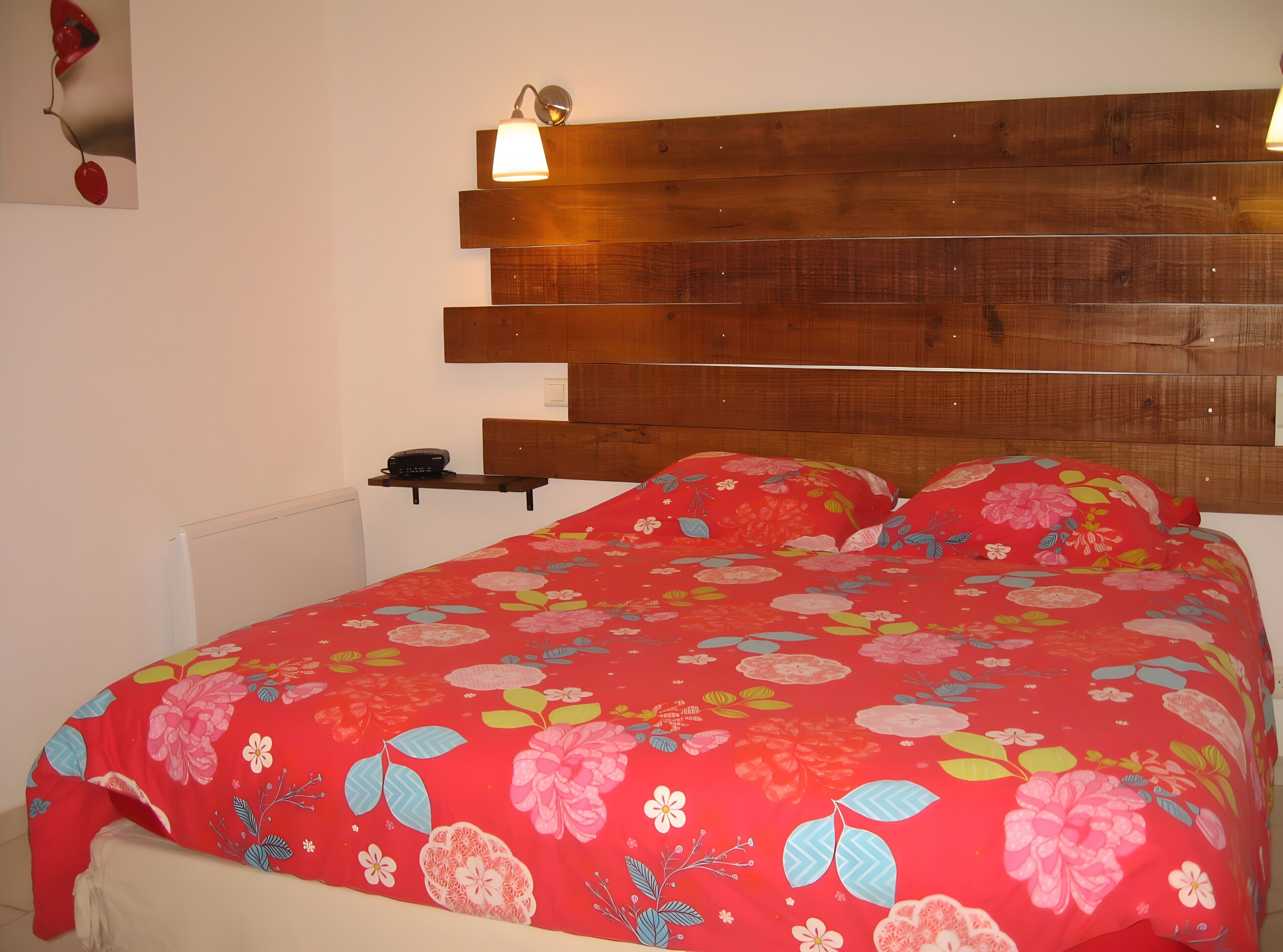 Ein Bett mit floralem Überwurf, ein hölzernes Kopfteil, ein Nachttisch mit Lampe und eine Wandleuchte.