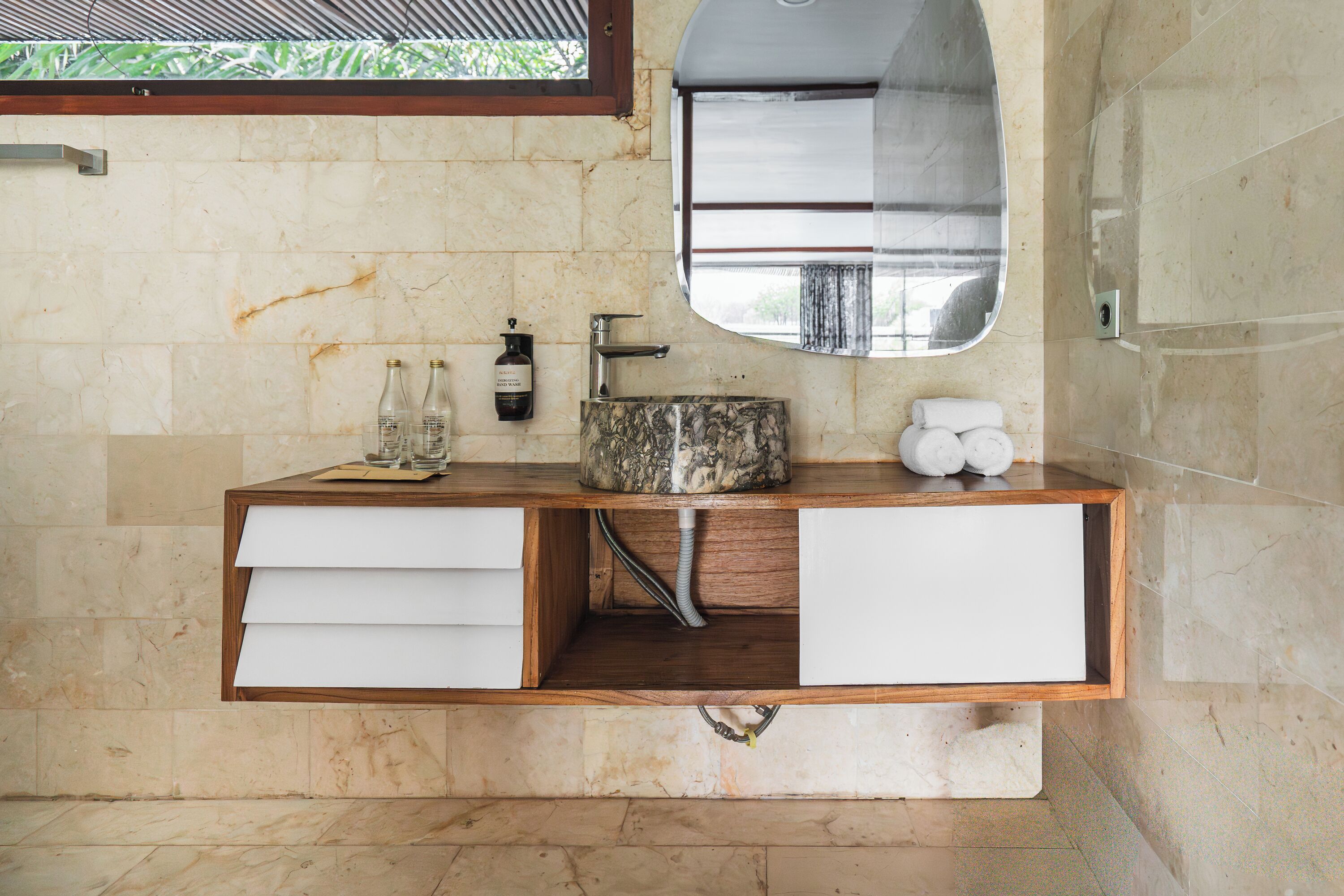 A bathroom with a marble-tiled wall, a wooden vanity with a sink, a mirror, and a window above the mirror.