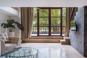 A spacious living room with a large window, a beige sofa, a glass coffee table, and a potted plant. - ONYX Park Resort Ubud (Ubud)