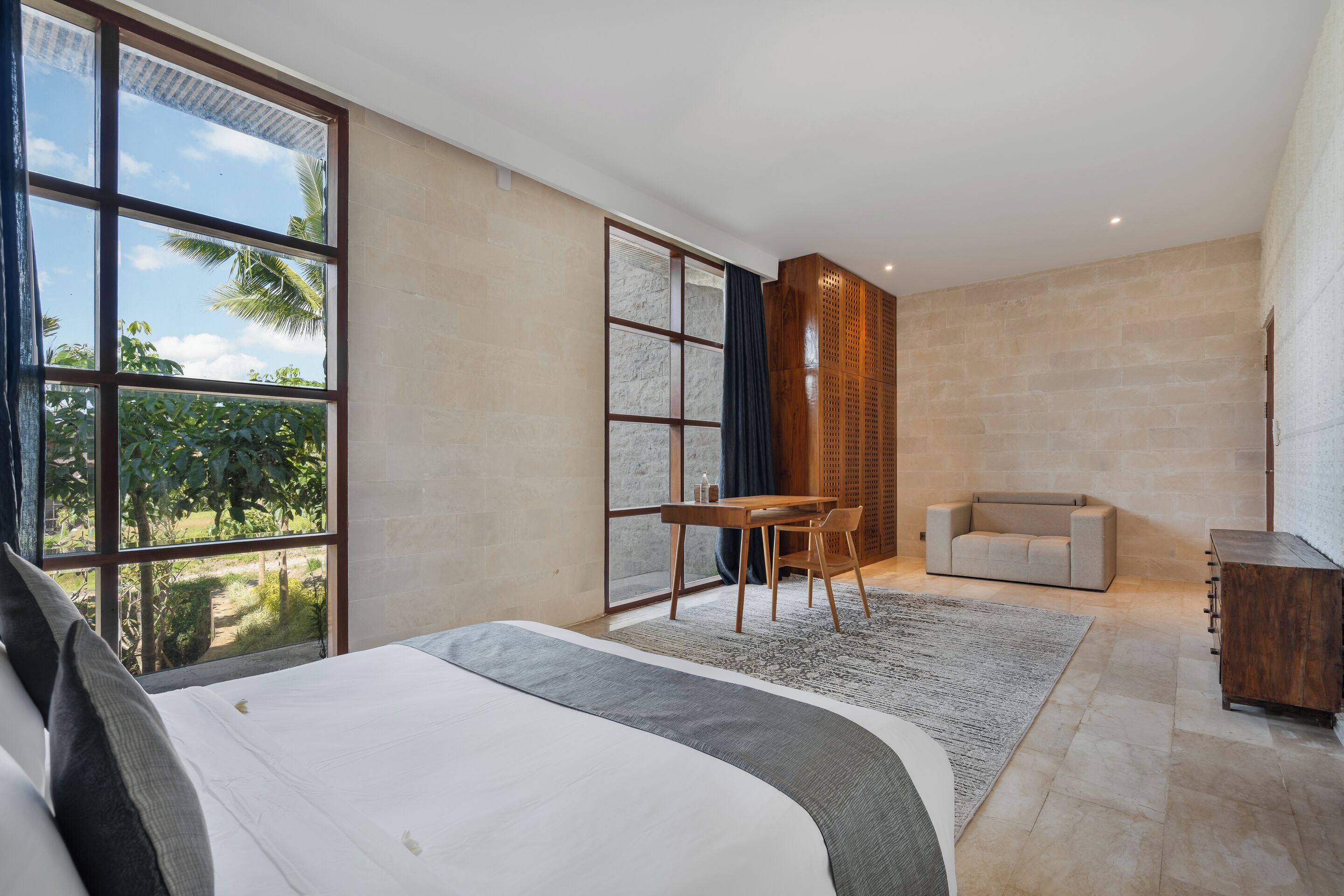 A modern bedroom with a large bed, a desk, a sofa, and a view of greenery through large windows.
