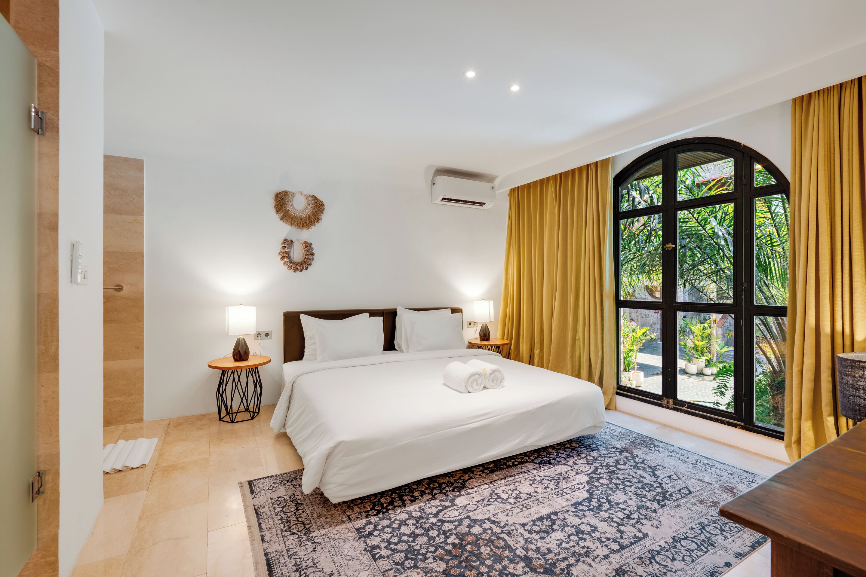 A hotel room with a large bed, two bedside tables, a window with curtains, and a view of greenery outside.