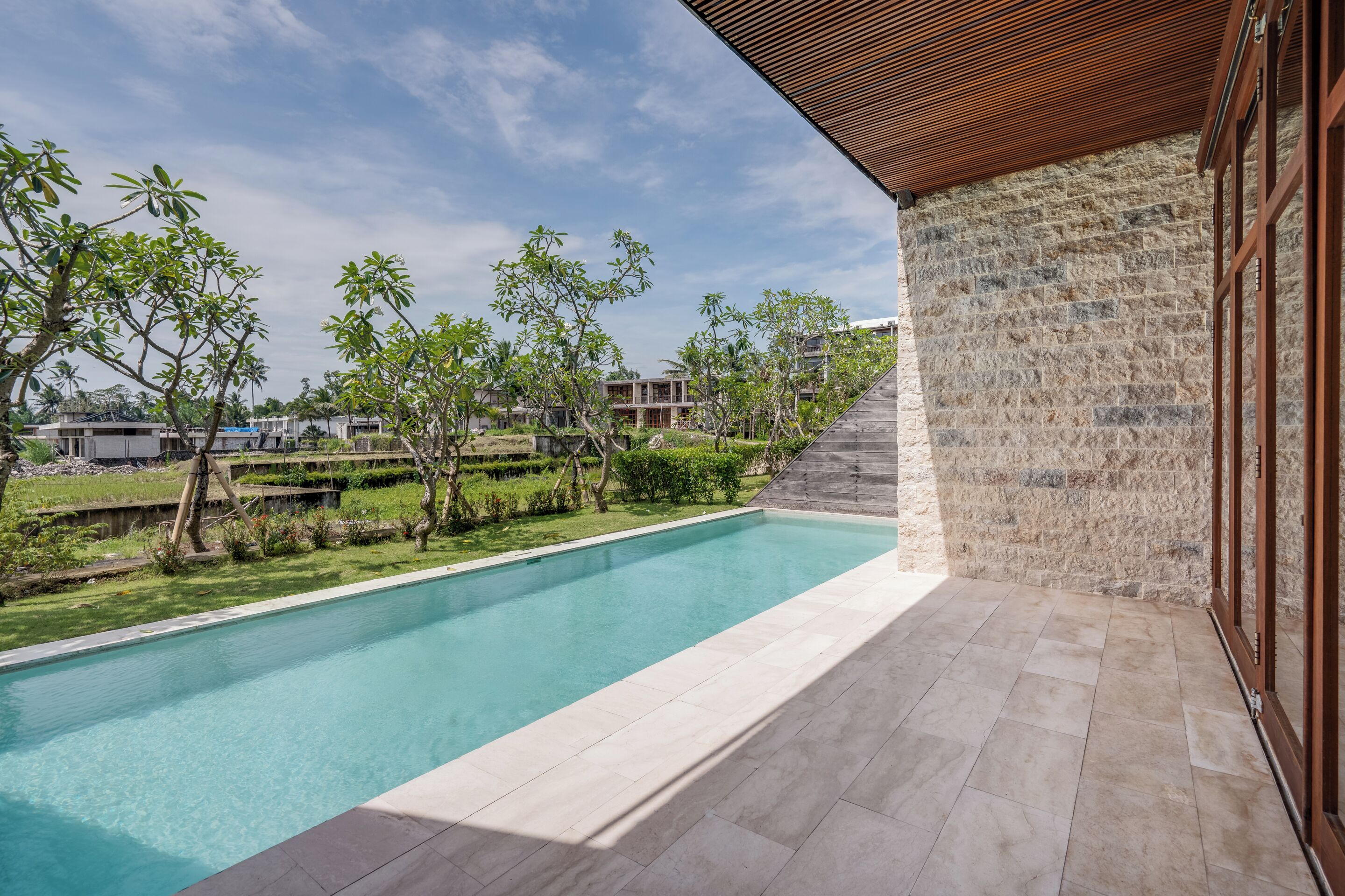 Une piscine bordée d’un mur en pierre et dotée d’un plafond en bois, entourée de verdure et surplombée par un ciel dégagé.
