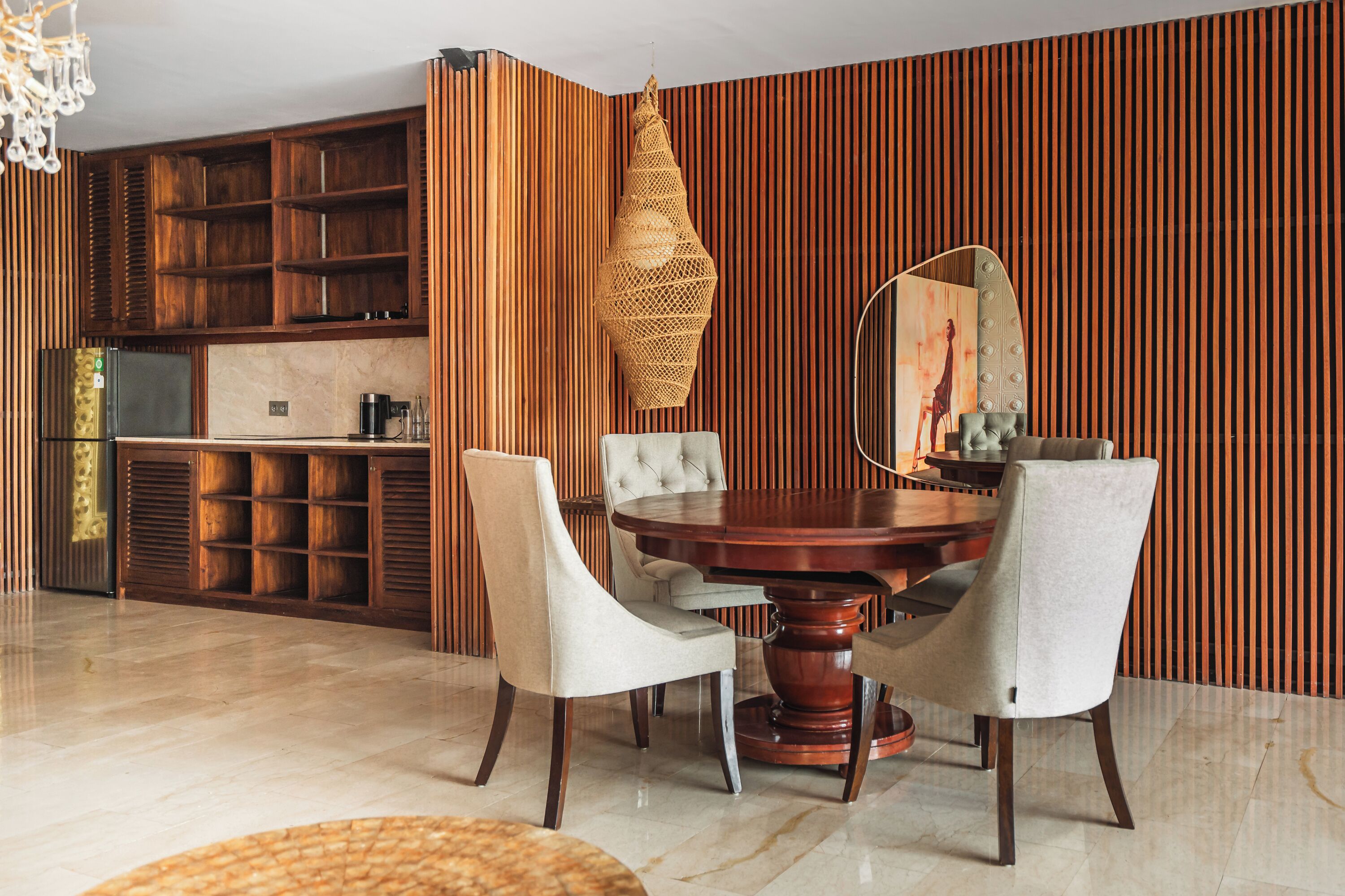 A dining area with a round wooden table, four upholstered chairs, a woven light fixture, and a kitchen in the background.