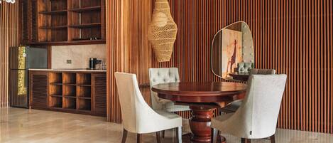 A dining area with a round wooden table, four upholstered chairs, a woven light fixture, and a kitchen in the background.