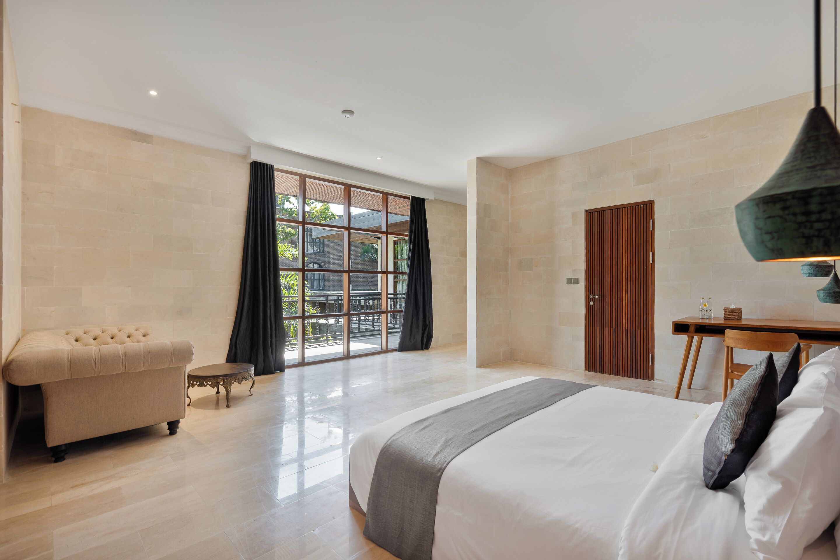 A modern hotel room with a large bed, a seating area, a dining table, and a view through large windows.