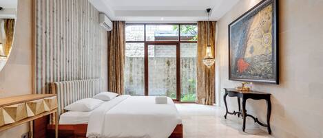 Quarto de hotel moderno com uma cama grande, uma mesinha de cabeceira de madeira, um quadro e vista para o verde através de uma porta de vidro.