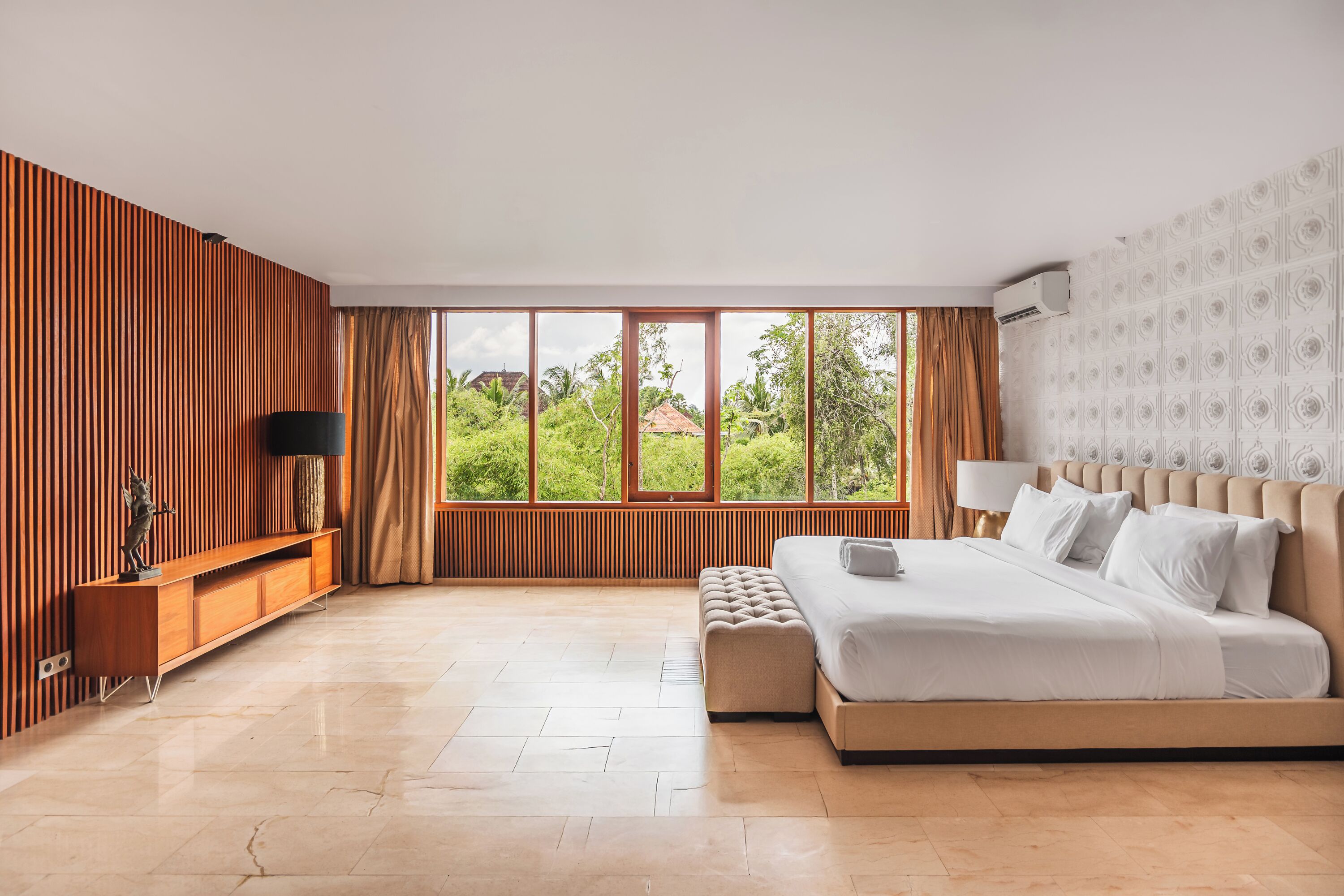 A modern hotel room with a large bed, a wooden panel wall, a view of greenery, and a window with curtains.