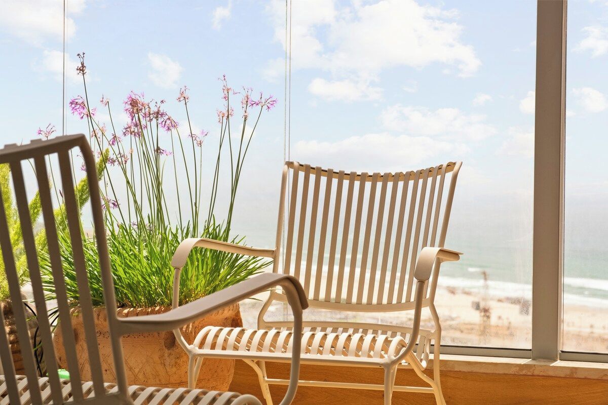Une chaise en osier blanche et une chaise en métal blanche sont placées face à une fenêtre donnant sur la plage et l’océan.