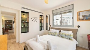 Habitación de hotel con cama, ventana, ventilador de techo y vista a edificios.