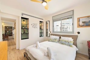 A hotel room with a bed, a window, a ceiling fan, and a view of buildings outside. - Bright Beachside Oasis | Steps to the Shore (Netanya)