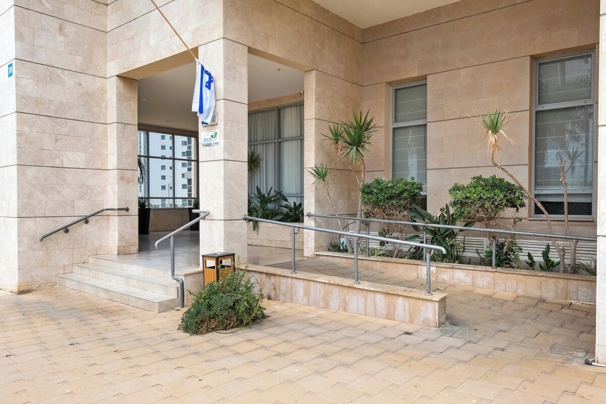 Entrada de prédio com rampa, corrimãos de metal e plantas em vasos.