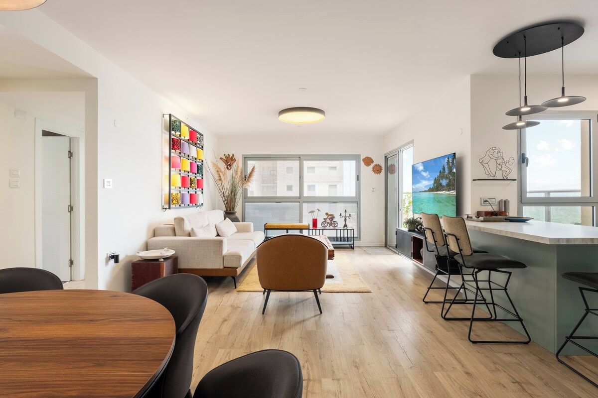 A modern living room with a sofa, coffee table, and chairs, a dining area with a wooden table and chairs, and a kitchen with bar stools.