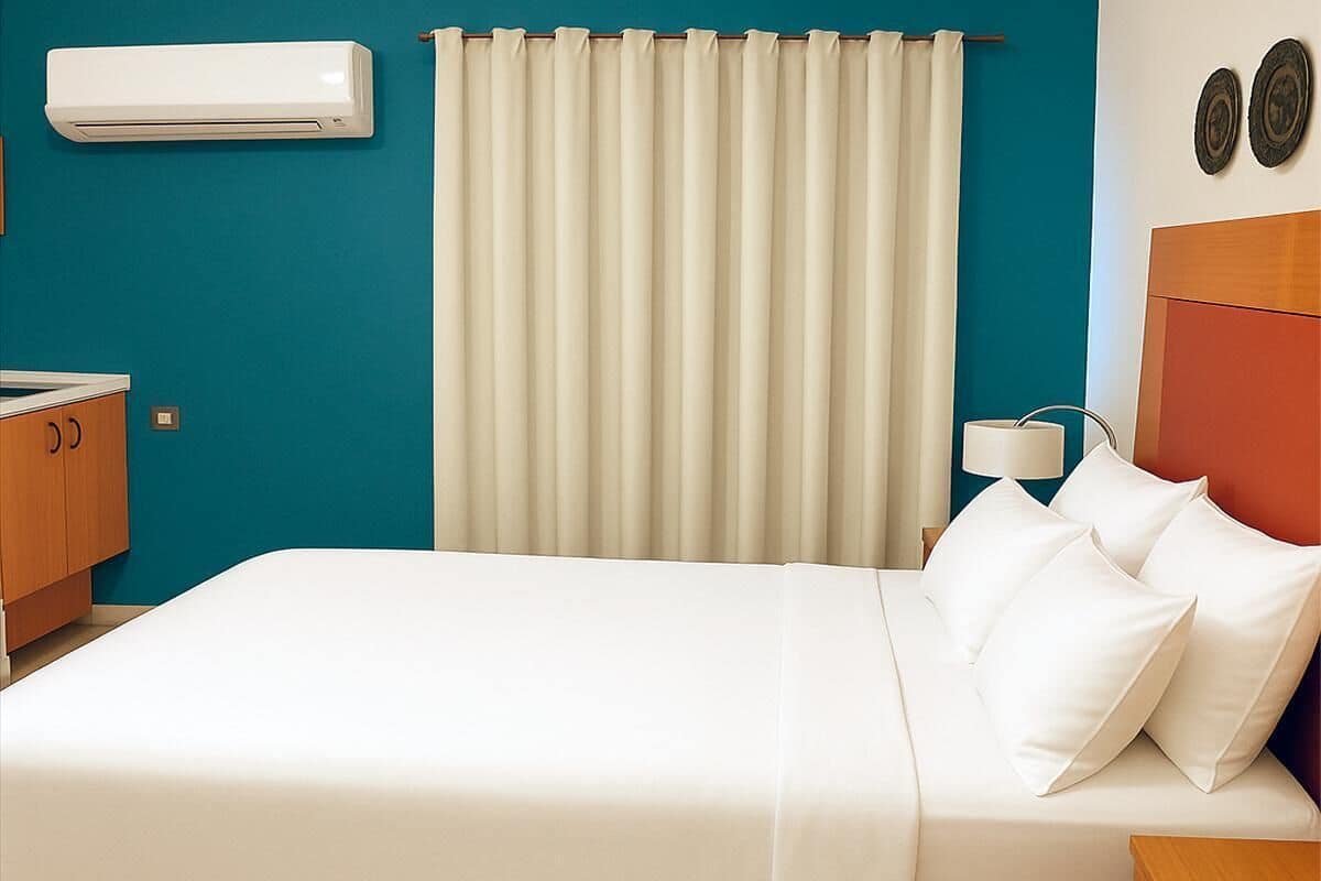Une chambre d’hôtel comprenant un lit, une table de chevet, une lampe, un climatiseur fixé au mur et un rideau.