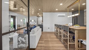 Una cocina moderna con armarios de madera, un comedor con una mesa larga y sillas, y una sala de estar con un sofá y un separador de vidrio.