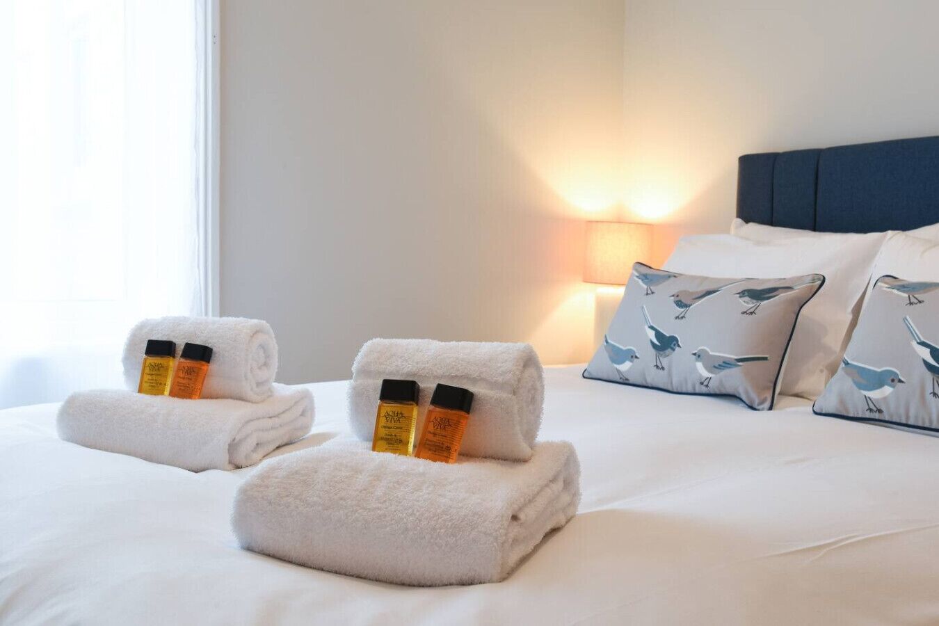 Ein ordentlich bezogenes Bett mit zwei aufgerollten Handtüchern, zwei Lotionflaschen, einer Nachttischlampe und Kissen mit Vogelmuster.