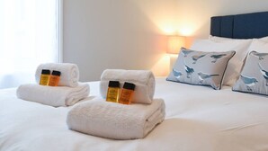 Ein ordentlich bezogenes Bett mit zwei aufgerollten Handtüchern, zwei Lotionflaschen, einer Nachttischlampe und Kissen mit Vogelmuster.
