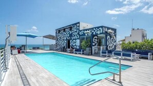Uma área de piscina com um edifício azul e branco estampado, céu claro e vista para o mar.