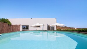 Une piscine avec une terrasse en bois, un bâtiment blanc doté d’une porte et de fenêtres, et deux parasols.