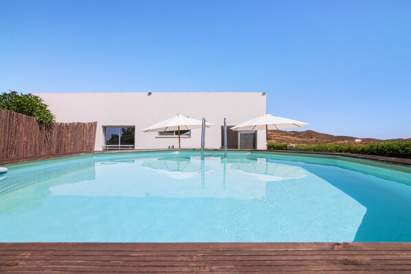 A swimming pool with a wooden deck, a white building with a door and windows, and two umbrellas.