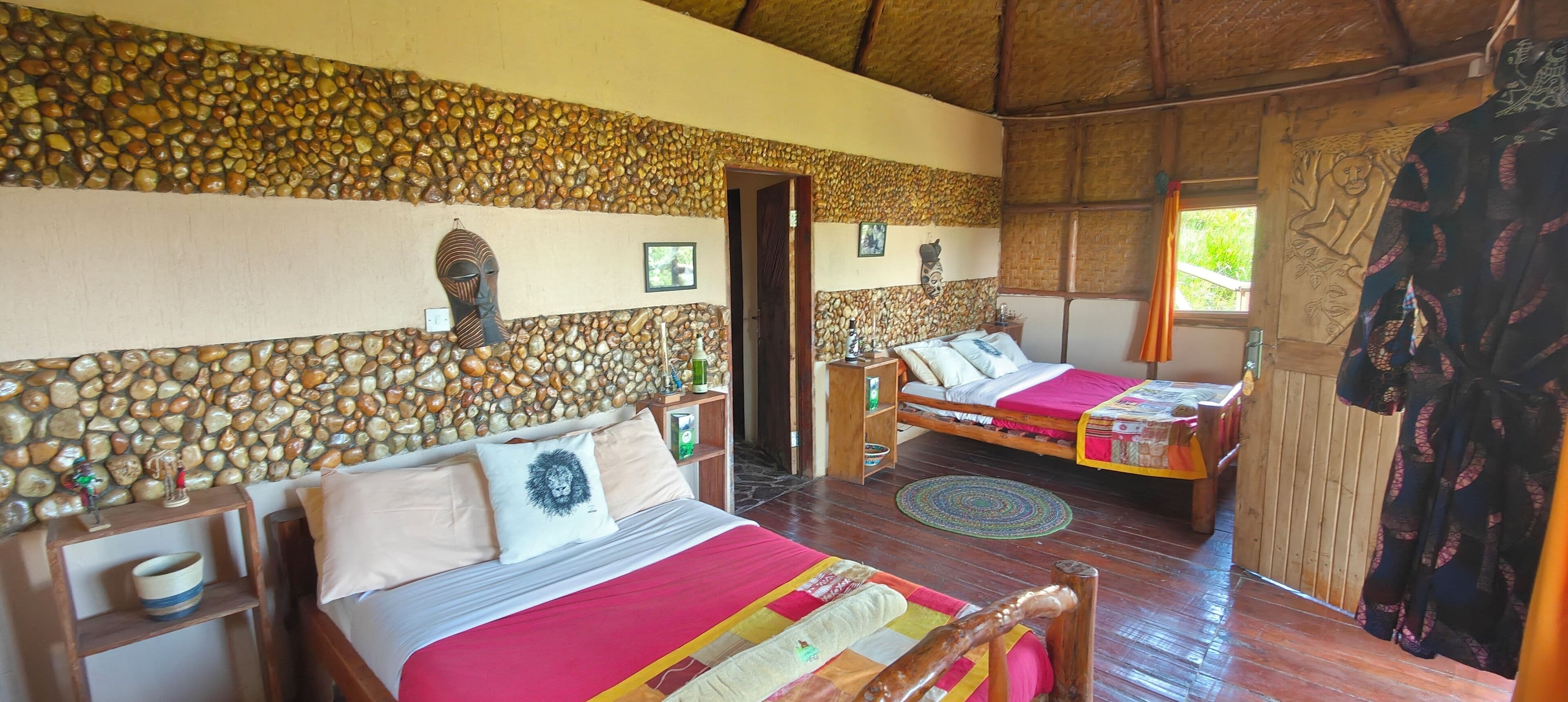 A room with two beds, a stone wall, wooden furniture, and a thatched roof.