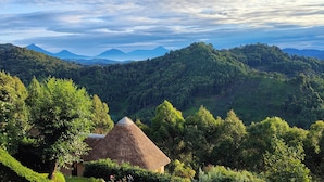 Ländliche Landschaft mit reetgedeckten Hütten, umgeben von üppiger Vegetation und Bergen im Hintergrund.