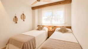 A room with two beds, a wooden ceiling fan, a window with a view of palm trees, and wall-mounted decorative items. - Residencial Marina Azul Beachfront (Denia)