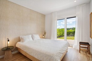 Ein Schlafzimmer mit einem Bett, einem Nachttisch, einer Lampe, einer Bank und einer großen Schiebeglastür, die auf einen Balkon führt.